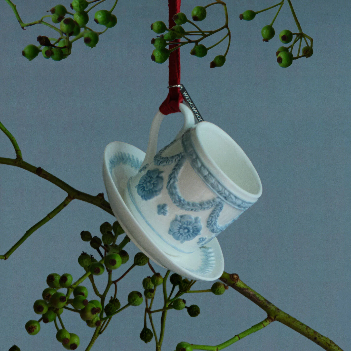Ritual Ornament White & Blue - A white and blue ceramic Wedgwood teacup and saucer ornament decorated with a blue floral pattern, hanging from a red ribbon on a tree branch with green berries against a blue-gray background.