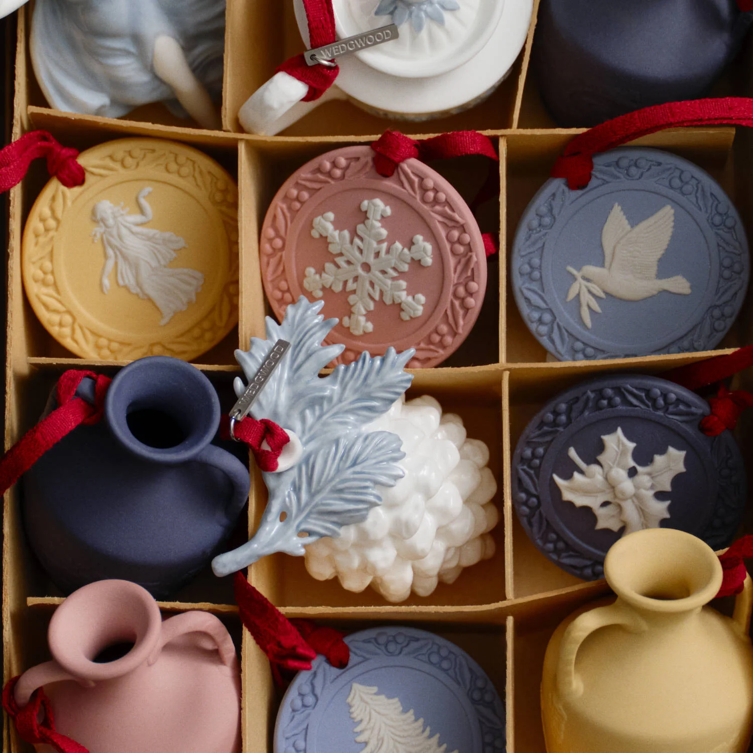 Dancing Hours Ornament Cane - A boxed assortment of small decorative Wedgwood jasperware ornaments and vases in muted pastel colors, each with a red ribbon loop and embossed holiday motifs. The set includes round medallions, a blue pitcher, a pink vase, a pale blue leaf ornament, a white grape cluster ornament, and other matching pieces arranged in a cardboard grid.