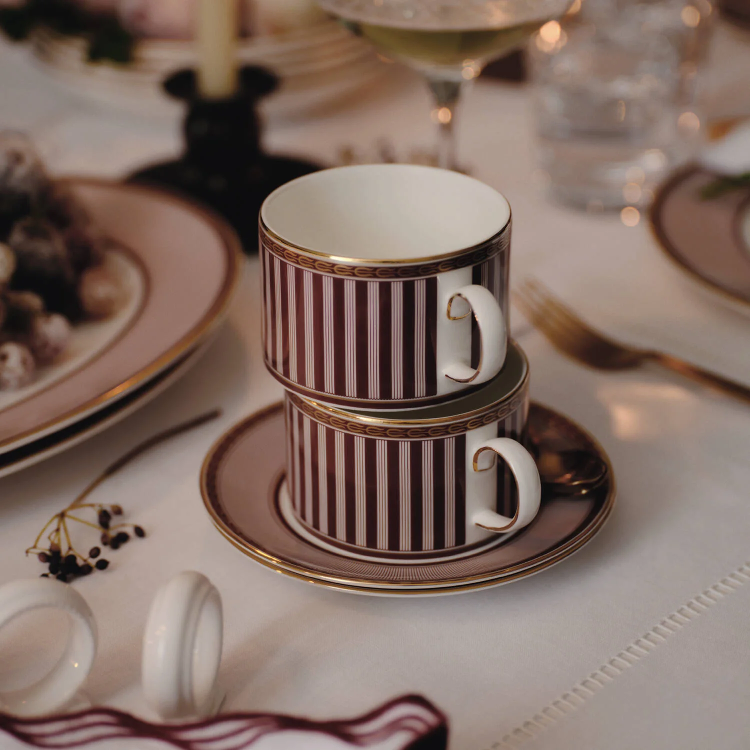 Splendour Red Teacup Saucer - Two stacked Red Splendour white ceramic teacups with burgundy vertical stripes and gold trim, resting on matching saucers on a white tablecloth at a dinner table.