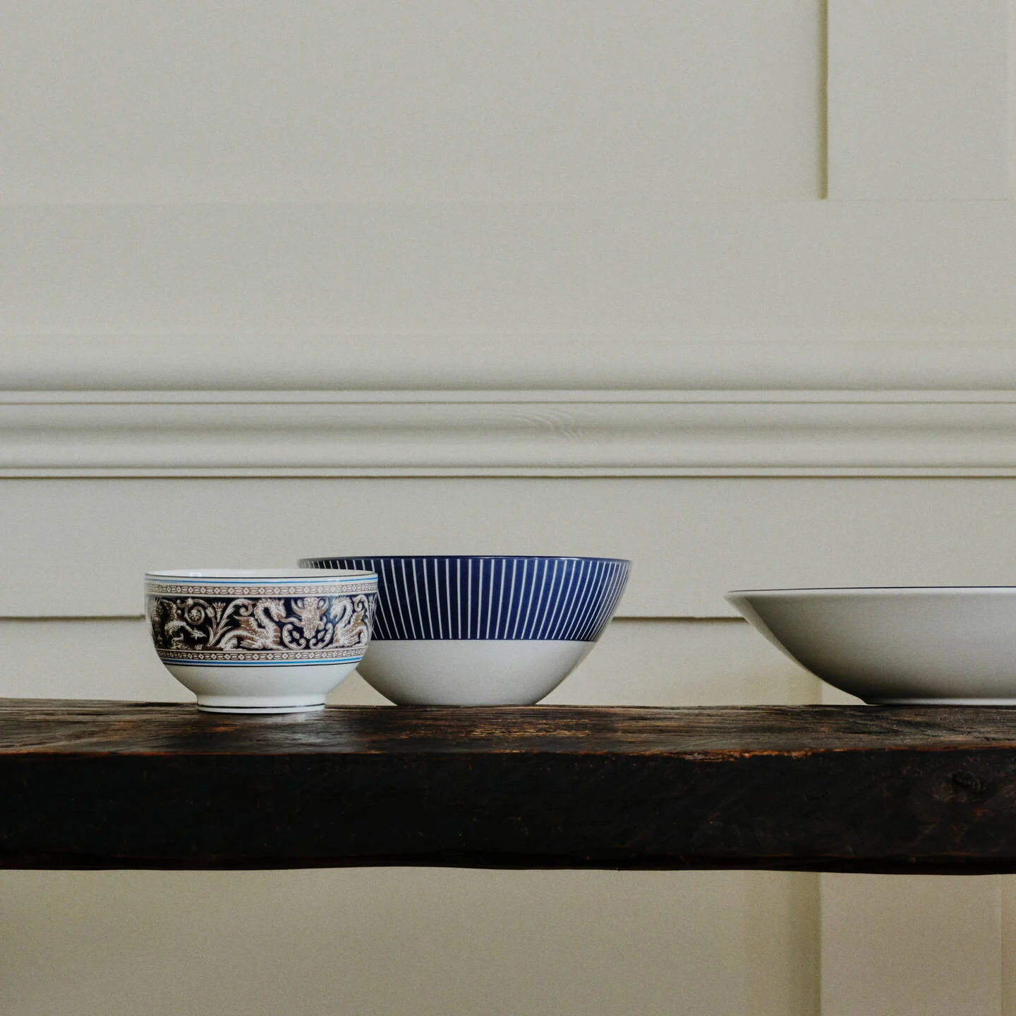 Florentine Navy Rice Bowl - Three ceramic bowls in Florentine Navy and Gio Blue pattern displayed on a dark wooden shelf against a cream paneled wall. The left bowl is white with a blue floral pattern, the middle bowl is white with blue vertical stripes, and a larger white bowl is partially visible on the right.