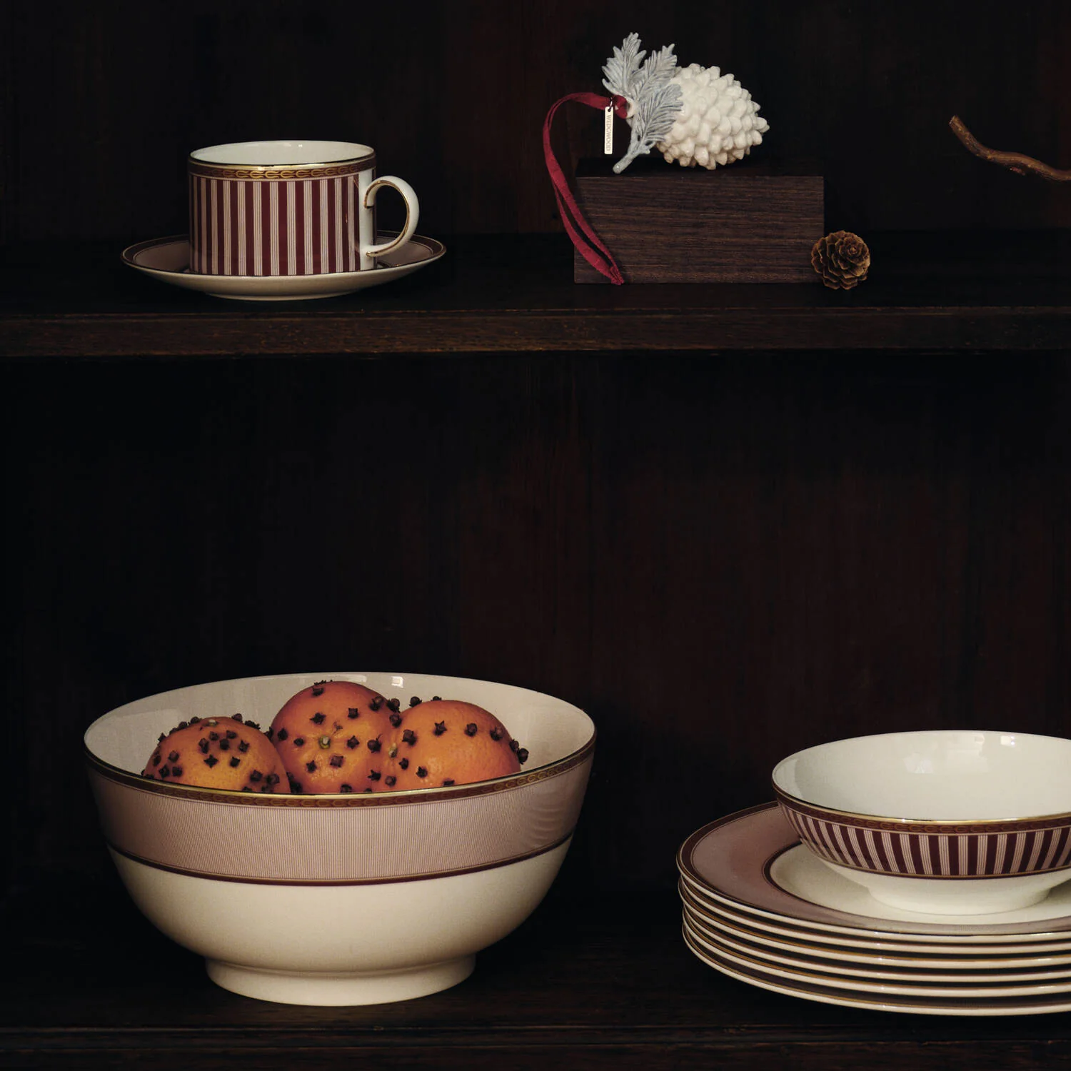 Splendour Red Serving Bowl - A set of elegant Red Splendour ceramic tableware displayed on a dark wooden shelf, including a striped cup and saucer, a large bowl with a beige band, a stack of plates, and a matching small bowl.