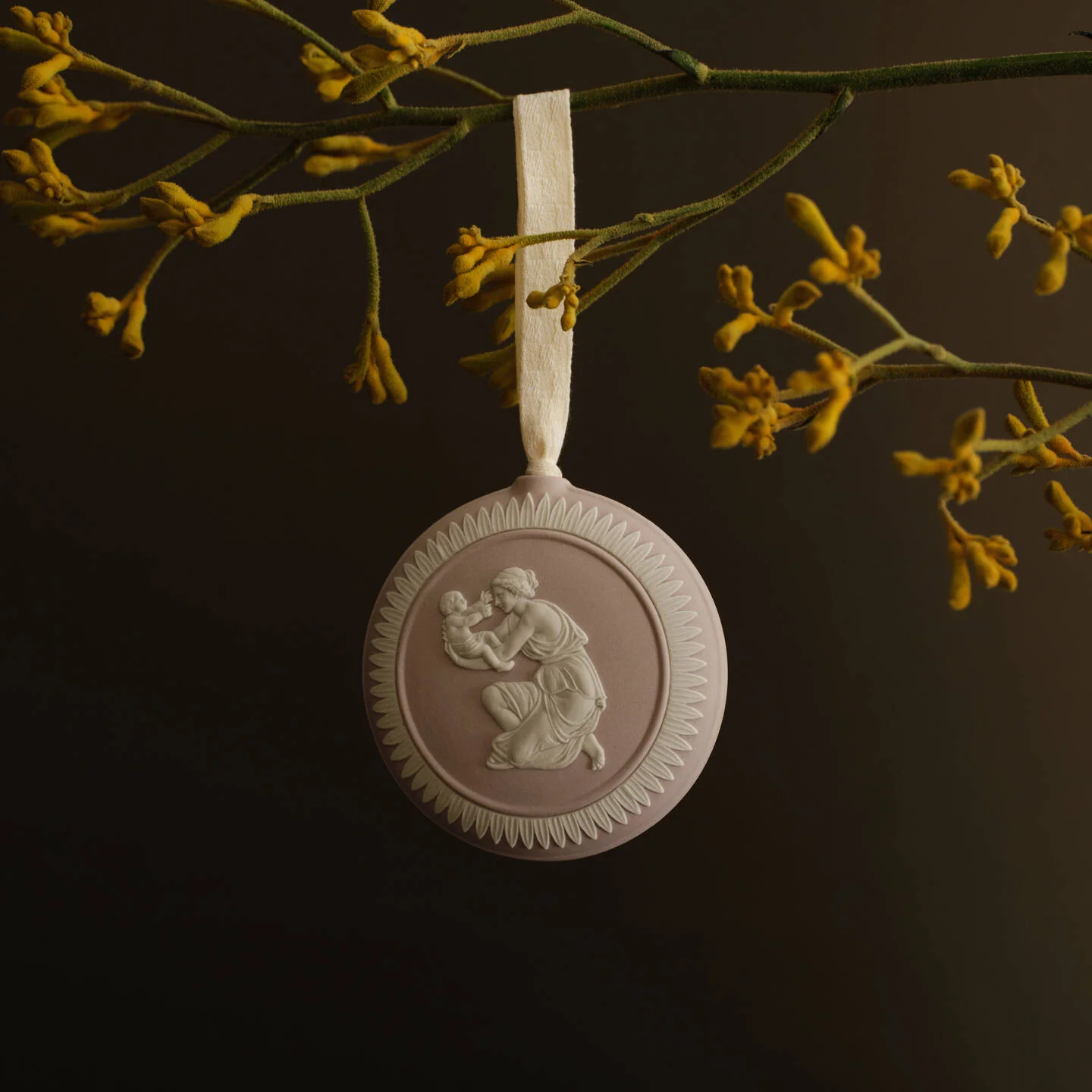 Mother Child Annual Ornament Mink - A round pale pink and white Wedgwood jasperware decorative hanging ornament with a relief figure design, suspended from a cream ribbon against a dark background with yellow flowers above.