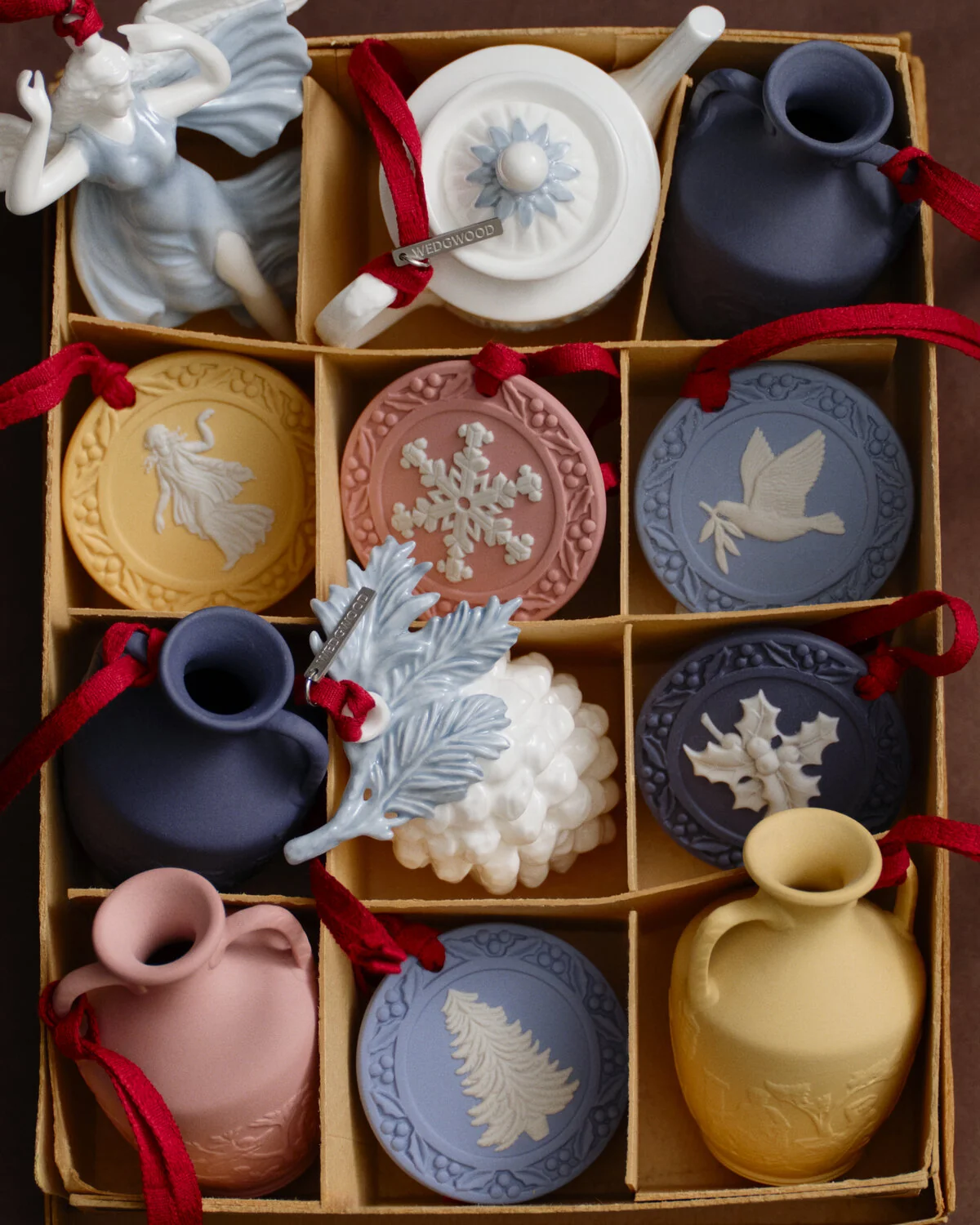 Holly Ornament Mercury Blue - A boxed assortment of Wedgwood decorative ceramic and Jasperware ornaments and small Portland vases arranged in a cardboard grid. The set includes round medallion-style ornaments with embossed winter motifs such as a snowflake, dove, holly, and tree, along with small vase-shaped pieces and a white figurine, all tied with red ribbons.