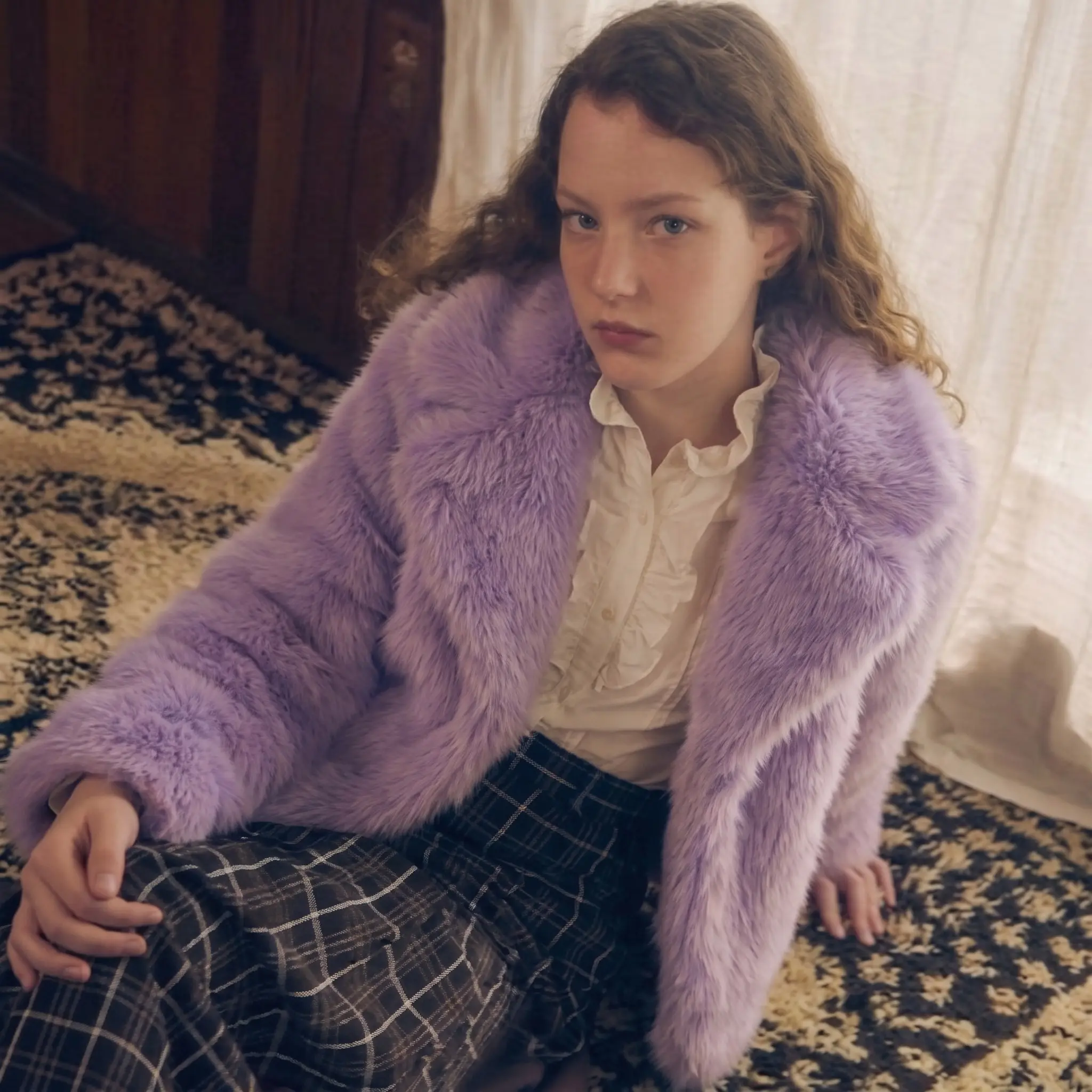 Woman wearing a lavender faux fur coat while relaxing in a bedroom