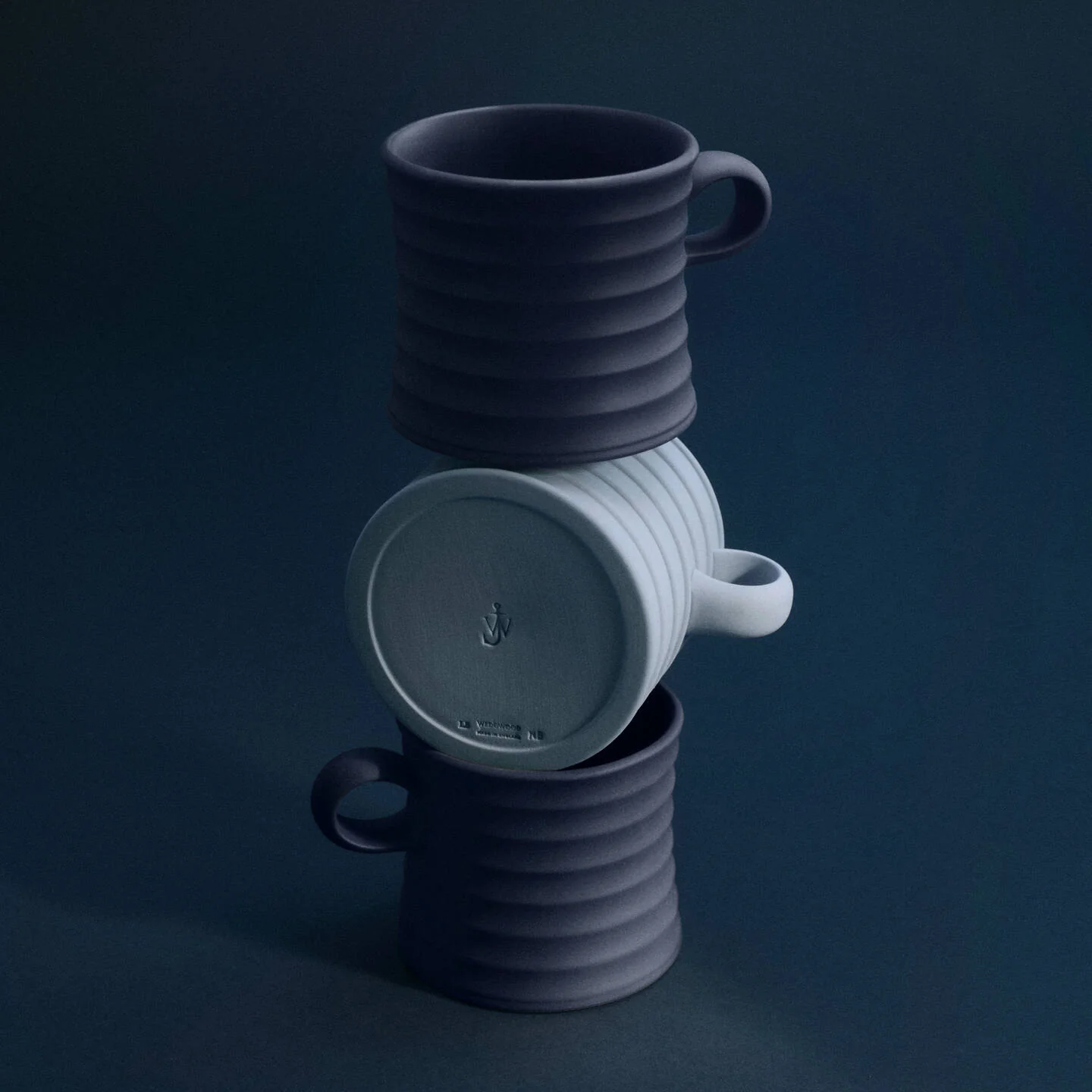 JW Anderson Mug Pair Mercury / Jasper Blue - Three ribbed JW Anderson jasperware mugs stacked in a tower on a dark navy background. The top and bottom mugs are dark charcoal gray, and the middle mug is a light gray.
