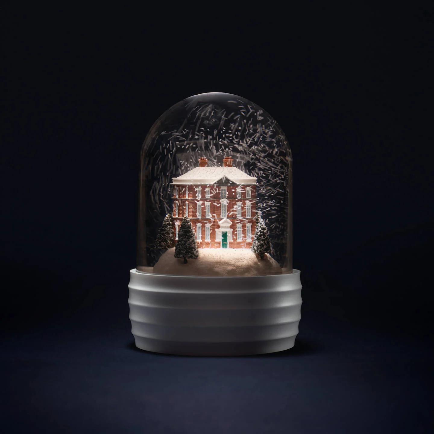 Snowglobe Wedgwood House - A decorative Wedgwood house glass dome snow globe on a ribbed white base, featuring a miniature red brick house surrounded by snow and trees, photographed against a very dark studio background.