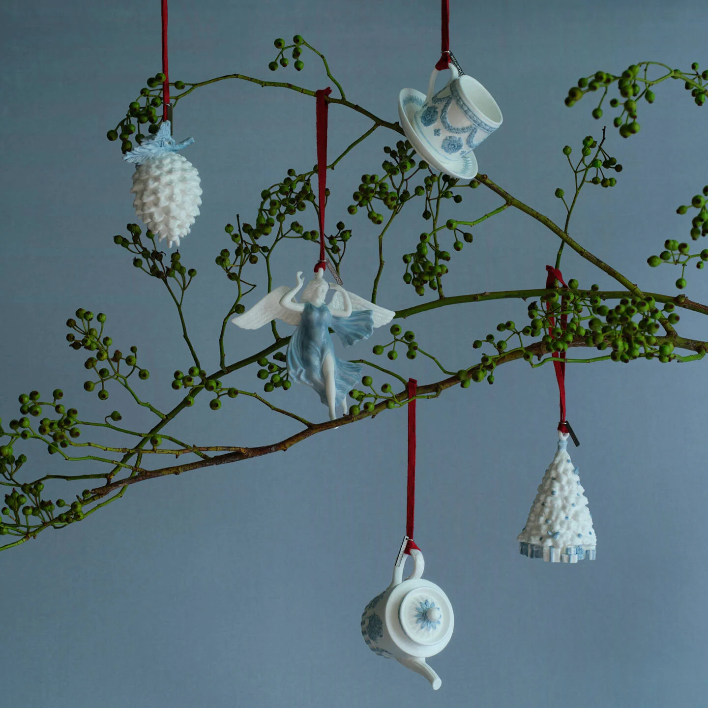 Love Ornament White & Blue - Five ceramic Wedgwood blue and white Christmas ornaments shaped like a pinecone, angel, teapot, teacup, and miniature Christmas tree, hanging from a decorative branch with red strings against a blue-gray background.