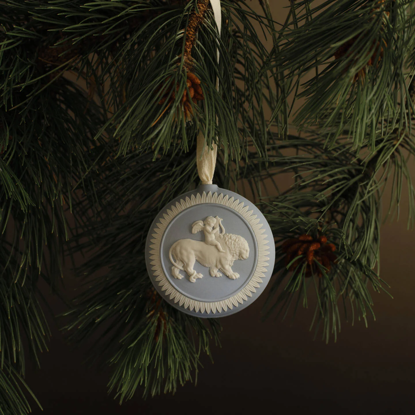 Lion Annual Ornament Light Blue - A round decorative hanging Wedgwood jasperware ornament with a lion and lamb motif, suspended from a green pine tree branch against a dark brown background.