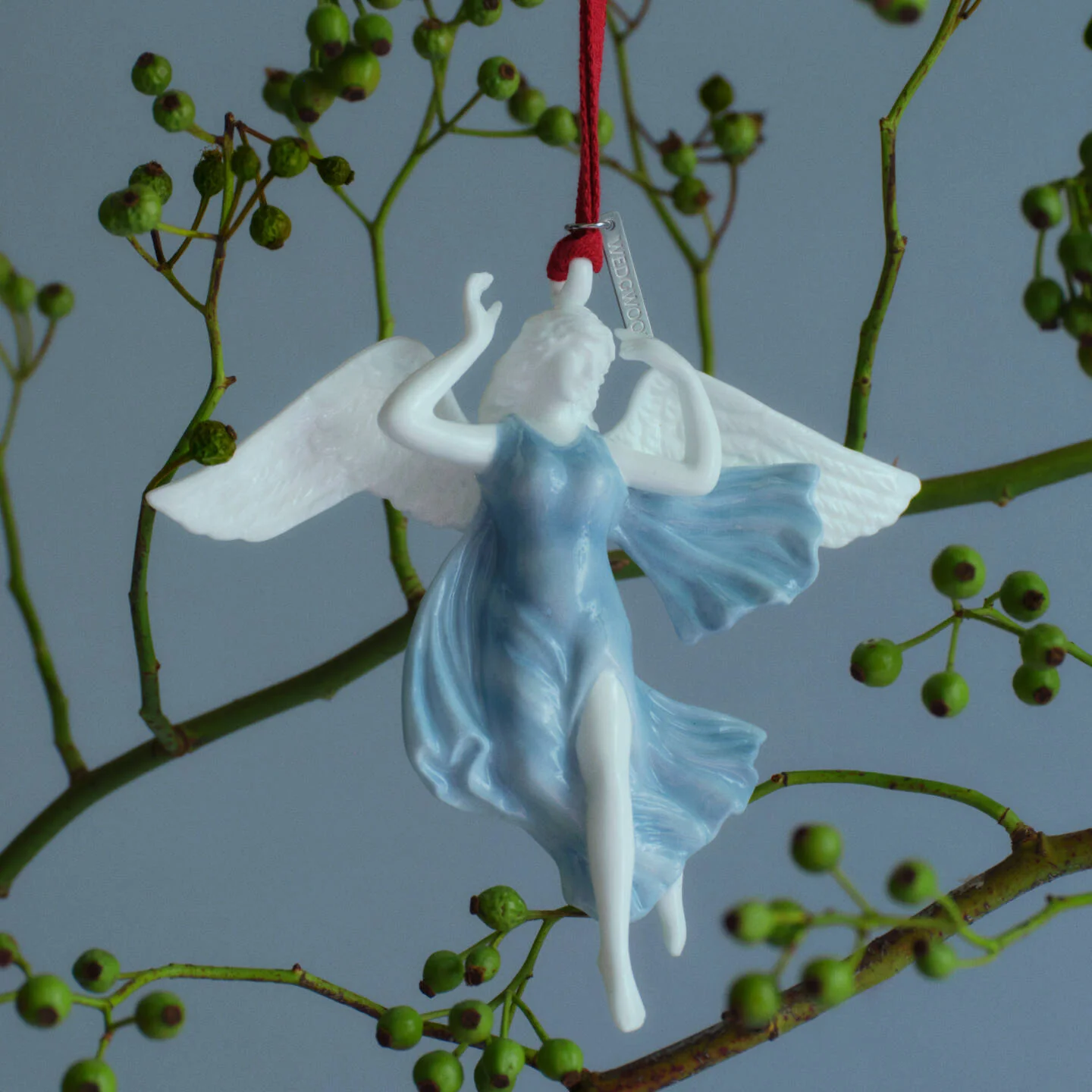 Peace Ornament White & Blue - A Wedgwood white and light blue angel Christmas ornament with outstretched wings, hanging from a red string against a gray background with green berry branches.