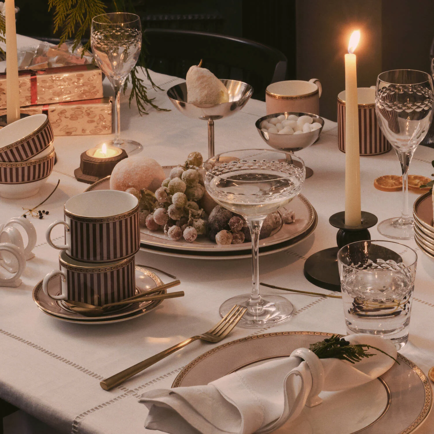Splendour Red Oval Platter - A warmly lit table setting with stacked Wedgwood Red Splendour ceramic mugs in burgundy and cream stripes, crystal-style drinking glasses, candlesticks, plates, napkins, and a centerpiece of grapes and fruit on a white tablecloth.